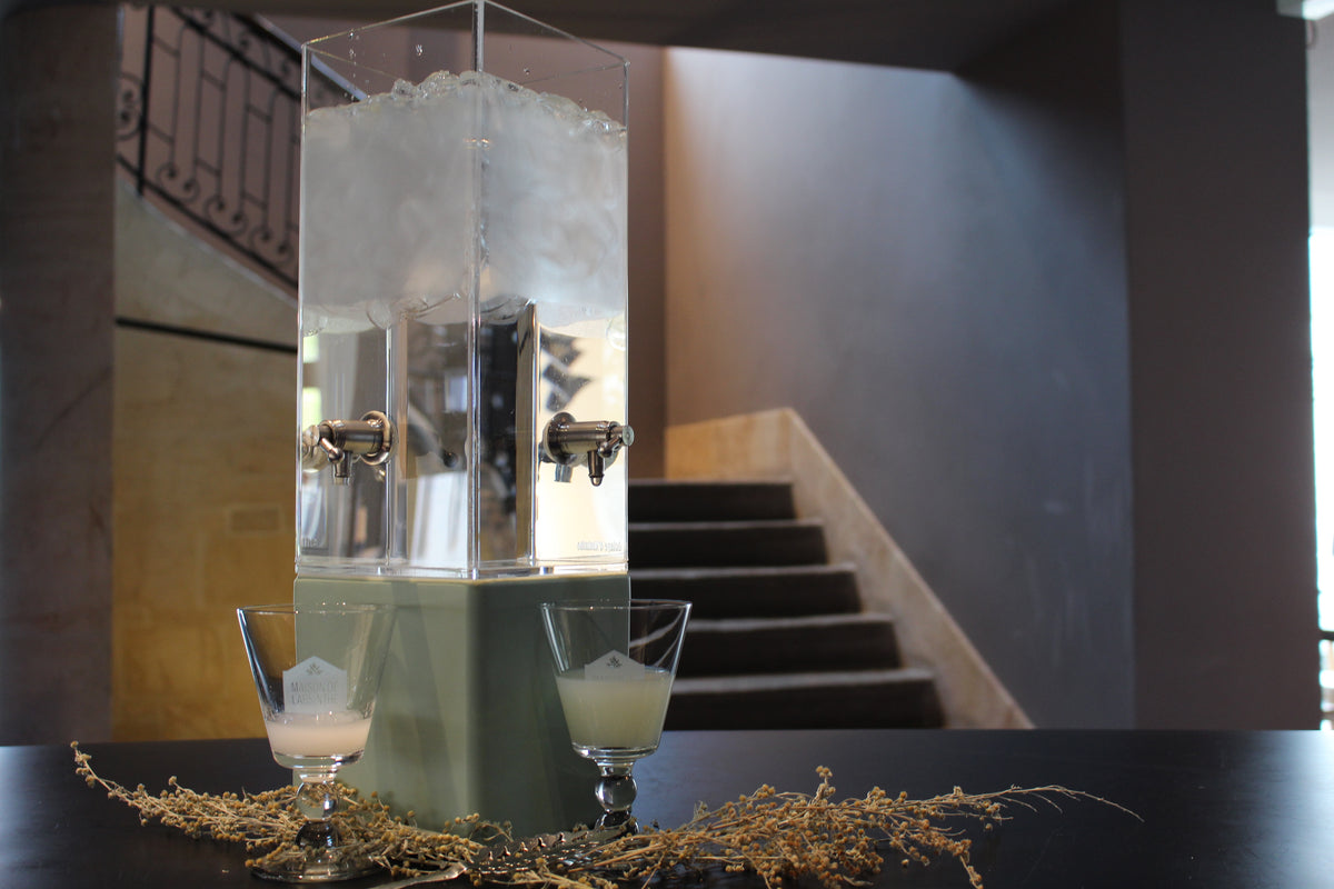 Two glasses of absinthe on stone steps beside a fountain, mid-ritual, water still dripping through the slotted spoons resting on their rims.