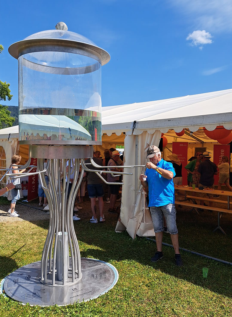 The museum's monumental Giant Absinthe Fountain standing on the grass at an open-air festival, a visitor in a blue shirt filling his glass at one of its taps, with white marquees and crowds behind under a summer sky.