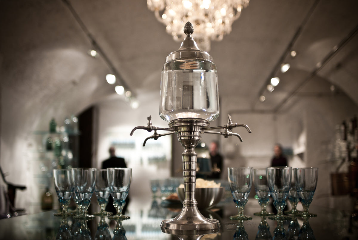 An antique four-tap absinthe fountain in pewter and glass stands on a dark polished bar between ranks of empty absinthe glasses, under a crystal chandelier in a white vaulted room.