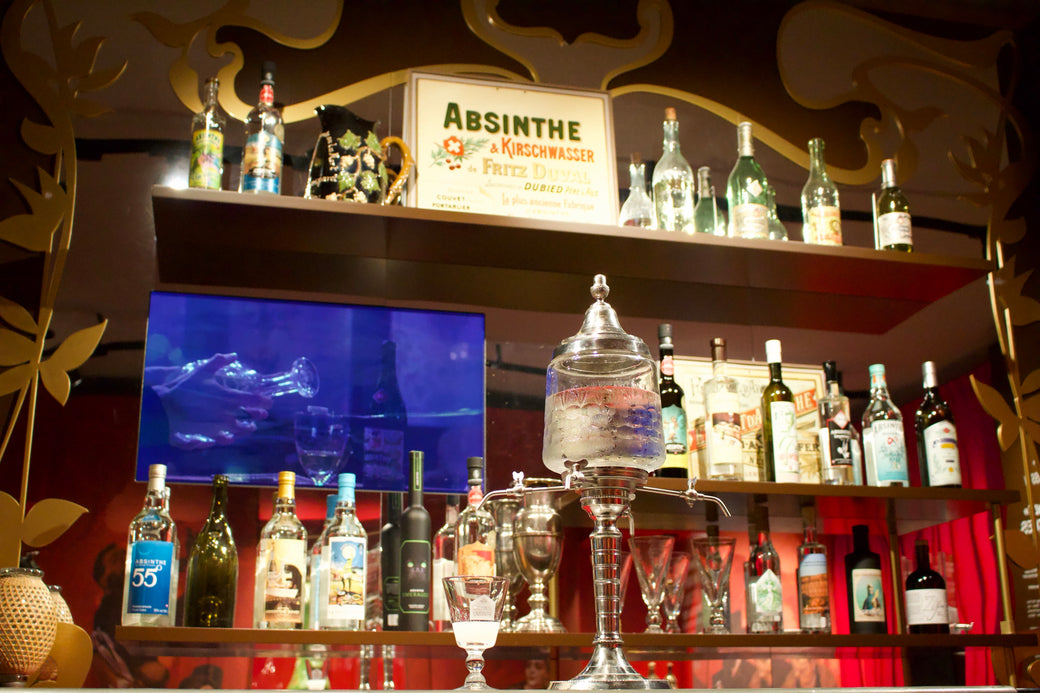 Traditional crystal absinthe fountain in the Belle Époque salon surrounded by vintage bottles and a Fritz Duval poster